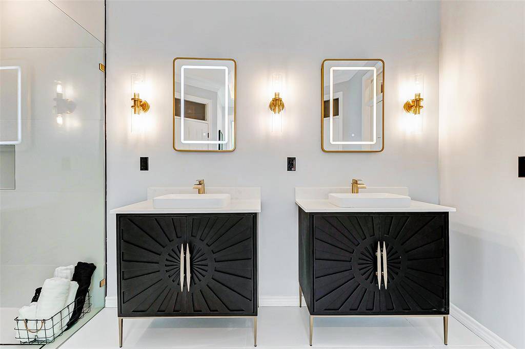 The dual vanities feature exclusive black cabinetry with Carrara quartz countertops and gold fixtures while the illuminated mirrors and sleek wall sconces add elegance.