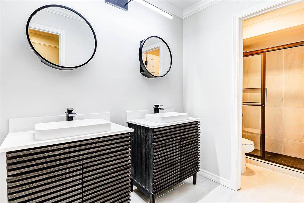 The modern en-suite bathroom features a sleek design with dual vanities with Carrara quartz countertops, round mirrors, and a spacious glass-enclosed shower, offering a stylish and functional space.