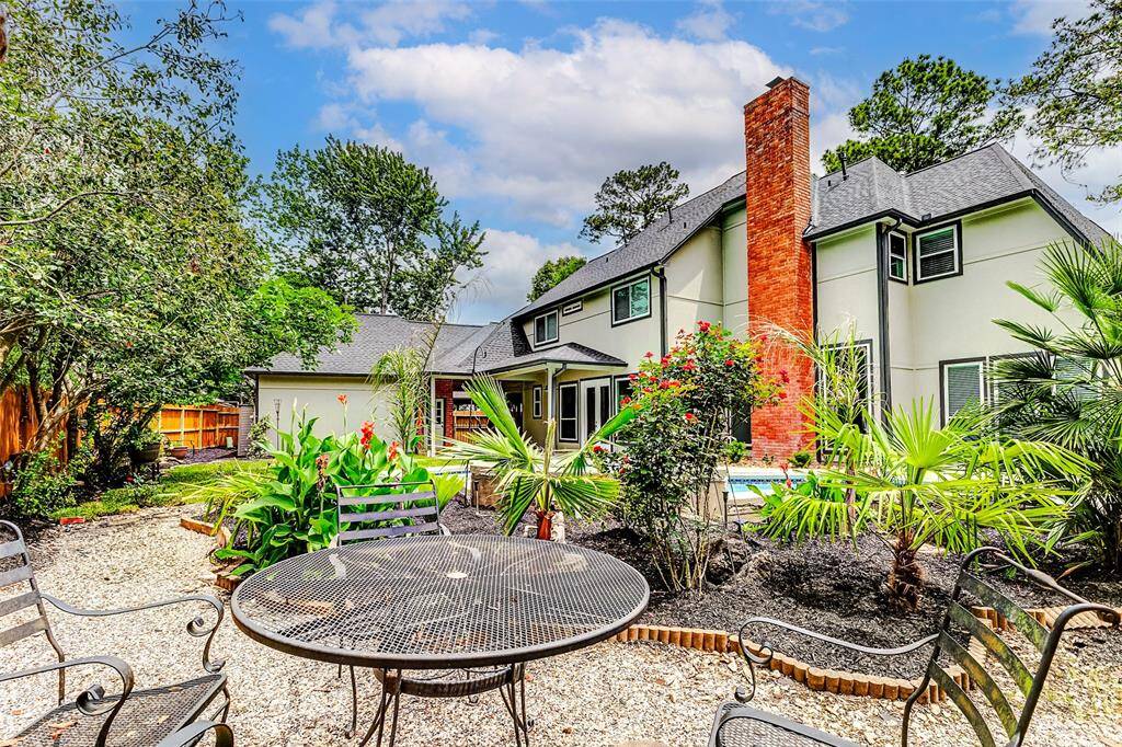 A charming backyard with flourishing landscaping, a cozy patio area with seating, and a view of a two-story house featuring a red brick chimney and multiple windows, creating a welcoming outdoor living space.