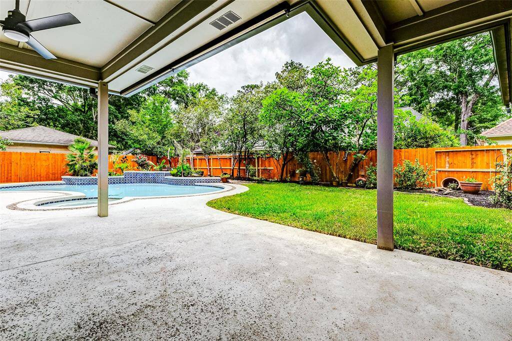 This backyard features a spacious covered patio with new concrete area around the pool and a new ceiling fan, overlooking a lush green lawn and a swimming pool with a raised spa.