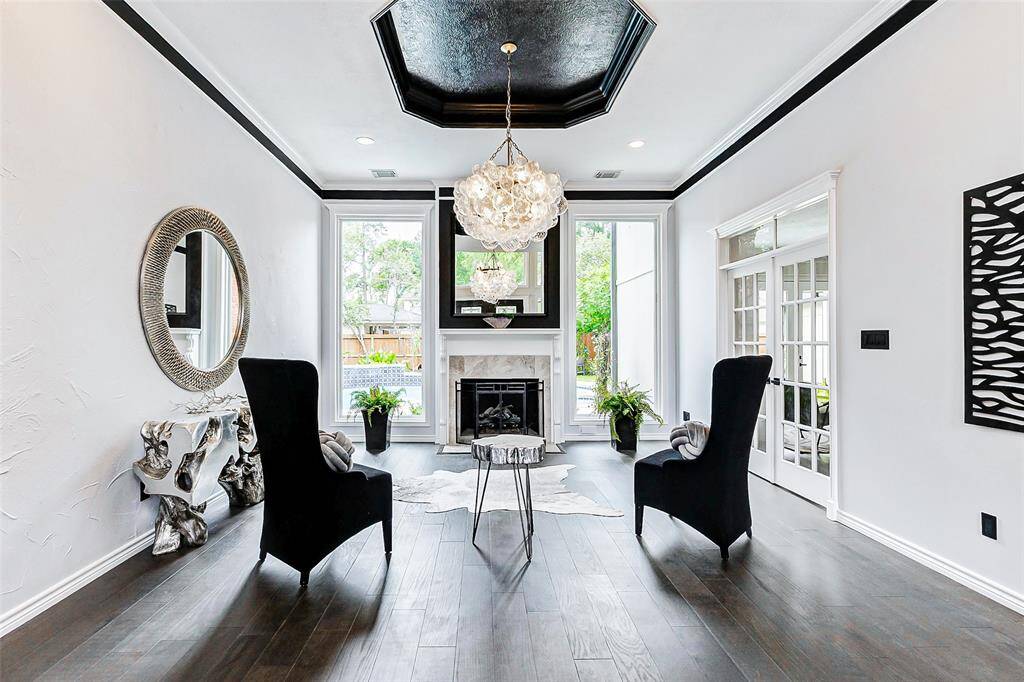 Bright, sophisticated living space with high ceilings, elegant chandelier, and large windows offering natural light. Features a central fireplace and paint accents creating a modern and inviting atmosphere.