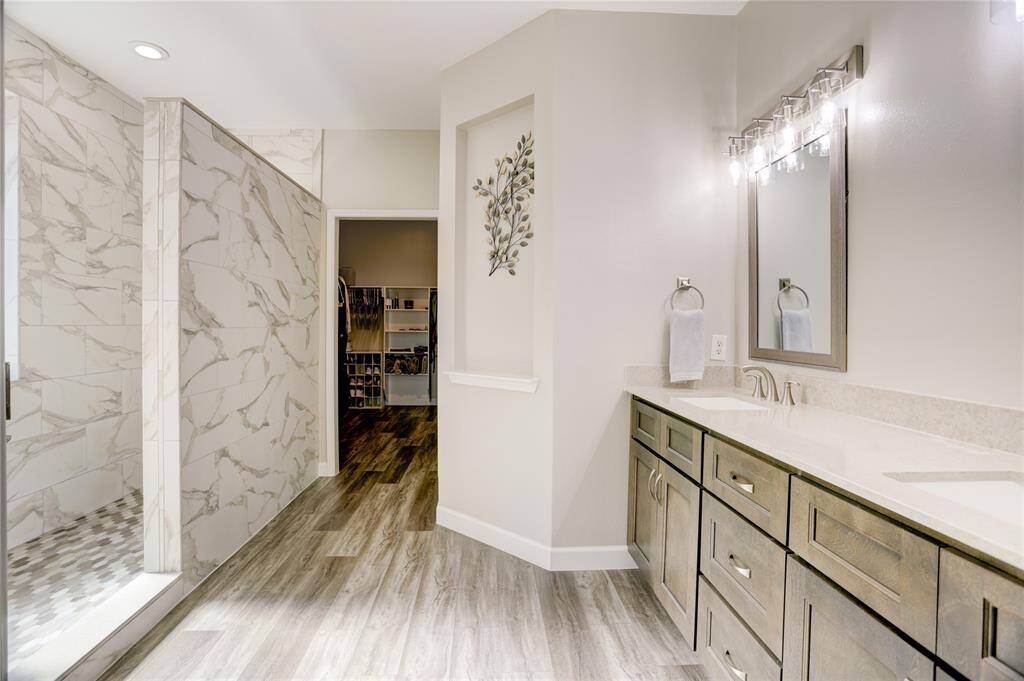 This bathroom features a spacious double vanity with elegant cabinetry and modern fixtures. It includes a large, tiled walk-in shower with a sleek, contemporary design. The neutral color palette and wood-look flooring create a warm, inviting atmosphere, leading into a roomy closet space.