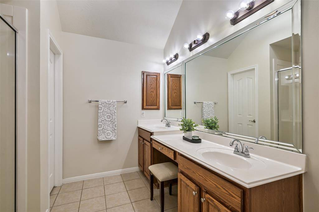 Primary bathroom offers dual sinks, a seated vanity area, an oversized mirror, and a separate shower, all accented by neutral tile and warm wood cabinetry.