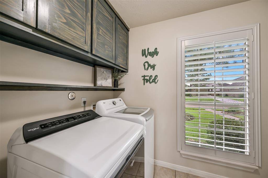 Spacious utility room with upper cabinetry, open shelving, and a large window with plantation shutters for natural light and front yard views.