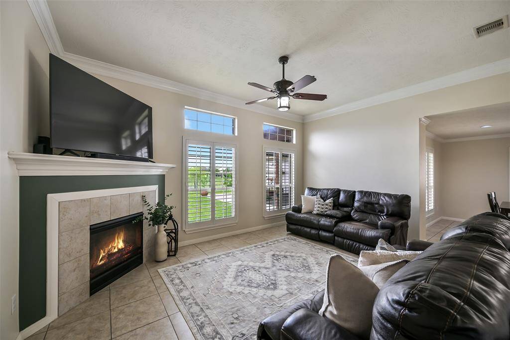 Another angle of the living room highlights the inviting gas fireplace with tile surround and custom mantle, tall ceilings with crown molding, and a wall of windows with transoms and plantation shutters that frame the peaceful lake view.