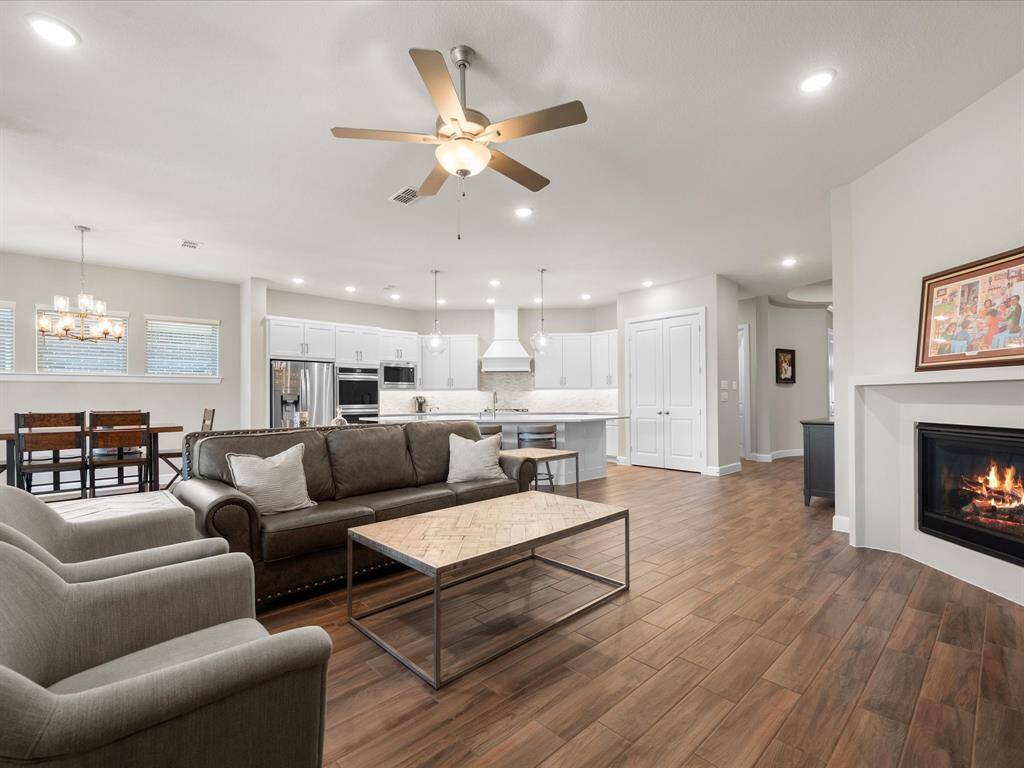 The spacious open-concept living room seamlessly connects to the kitchen and dining area, anchored by a cozy gas fireplace and rich wood-look tile throughout. The kitchen shines with white cabinetry, a large center island, stainless steel appliances, and pendant lighting—perfect for both entertaining and everyday living. Ample natural light pours in through the windows, creating an inviting and airy ambiance.