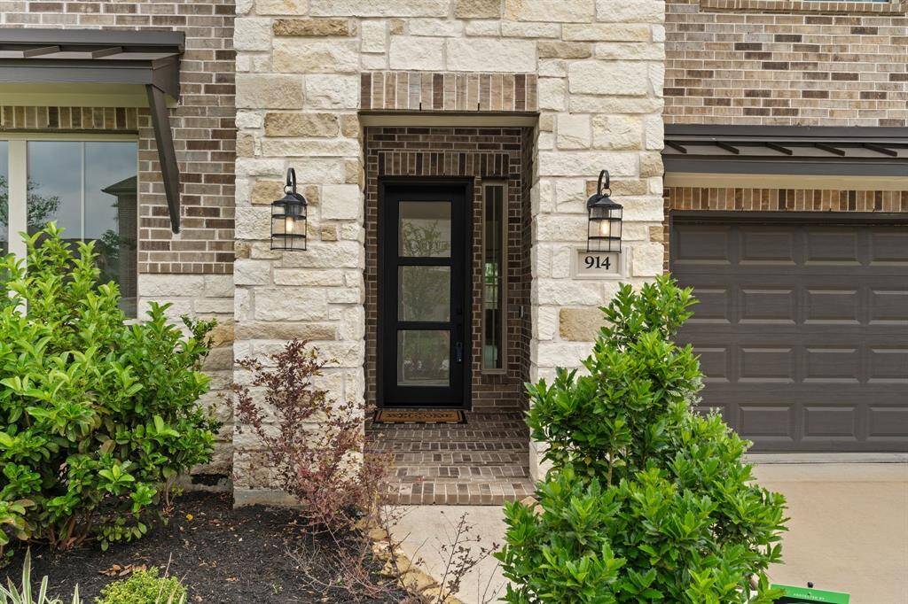 This photo showcases a modern home entrance with a blend of stone and brick exterior. The front door is sleek and dark, flanked by stylish outdoor lanterns. Well-maintained shrubs enhance the welcoming curb appeal.