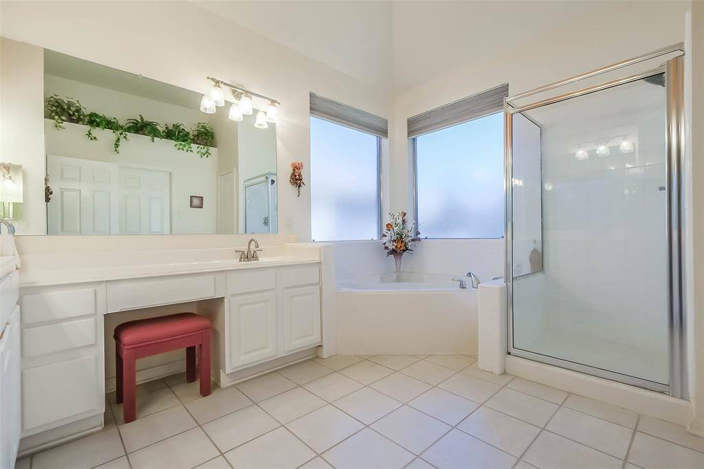 This bright, primary bathroom features a large vanity with ample storage, a comfortable jetted soaking tub, a separate glass-enclosed shower, and elegant tile flooring. Frosted windows provide privacy and plenty of natural light.
