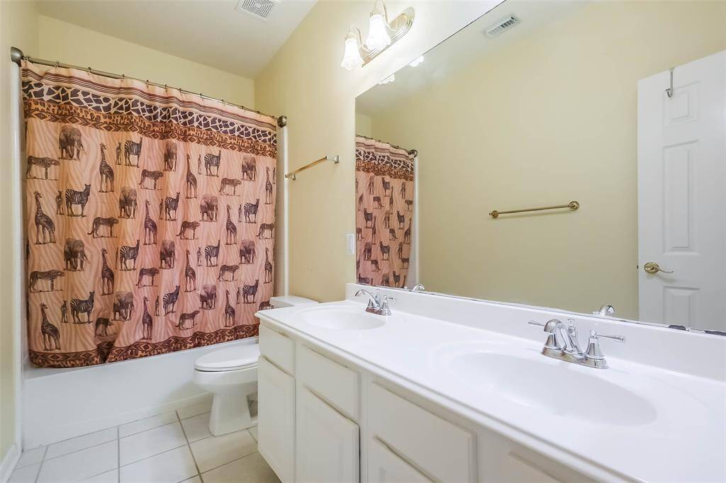 Full bathroom features a double-sink vanity with bright lighting and a large mirror. It has a shower-tub combination and is complemented by light walls and tile flooring, creating a clean and inviting atmosphere.