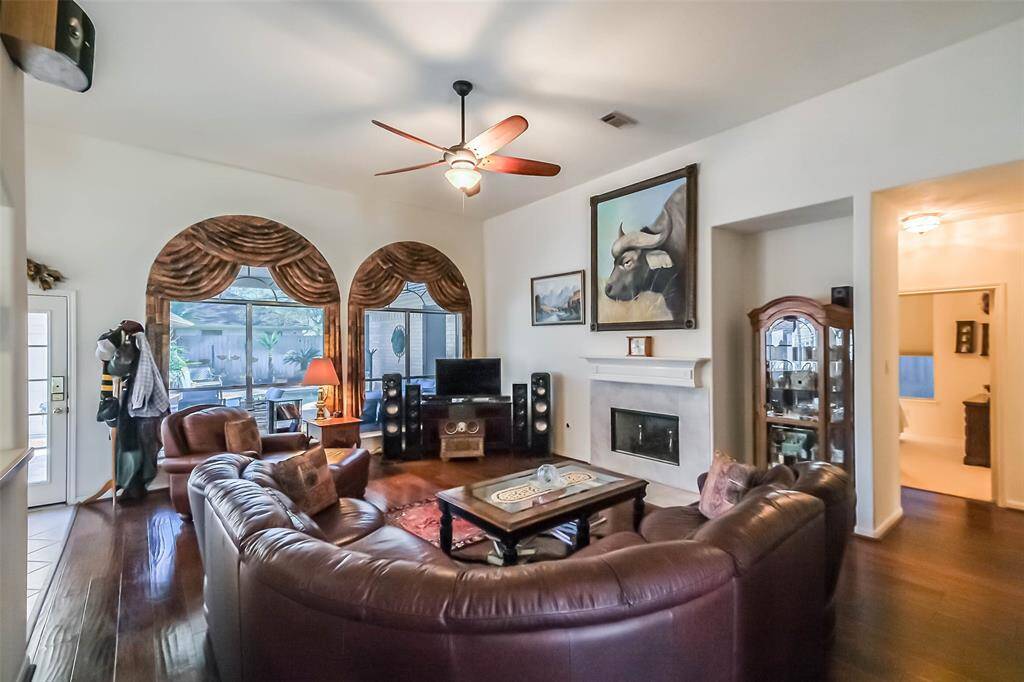 Spacious living room with high ceilings, featuring a cozy fireplace, elegant wood flooring, and abundant natural light from large windows. The room includes a stylish ceiling fan and is tastefully decorated with artwork and a comfortable leather sectional.