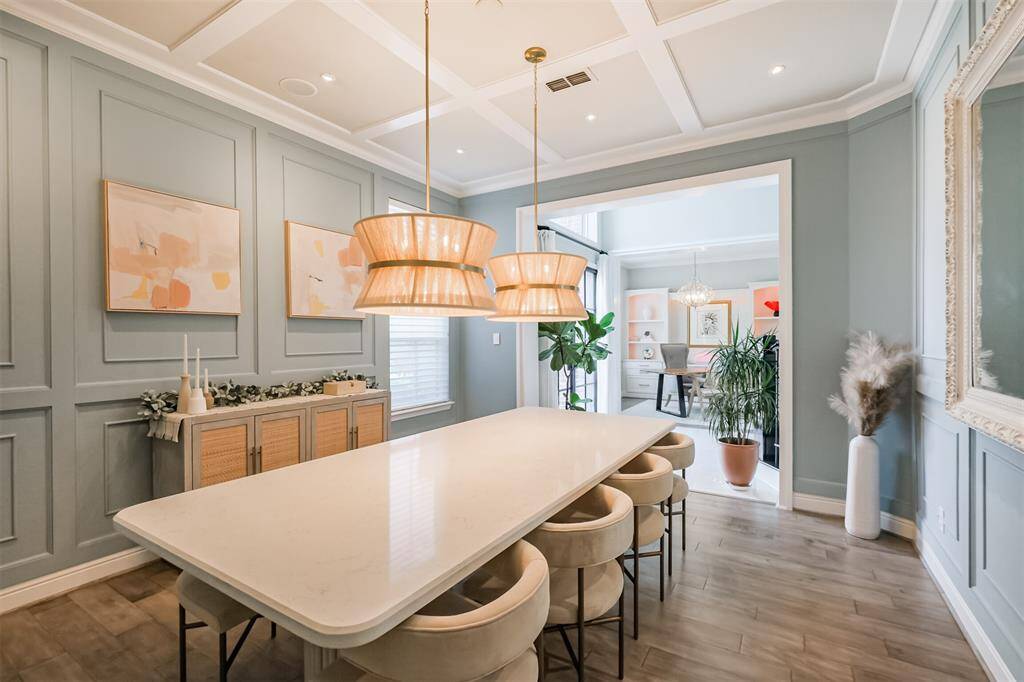 This dining room exudes timeless elegance with custom wainscoting that adds texture and sophistication to the walls, while a coffered ceiling introduces architectural depth and visual interest overhead. Two statement pendant lights, suspended within the coffered ceiling, serve as striking focal points, casting a warm glow that enhances the room's inviting ambiance.