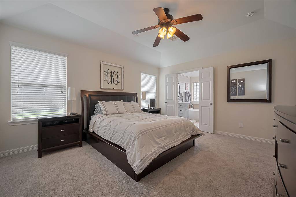 This inviting primary bedroom features two windows for natural light, a ceiling fan, plush carpet, and an en suite bathroom with double doors, creating a cozy and functional space.
