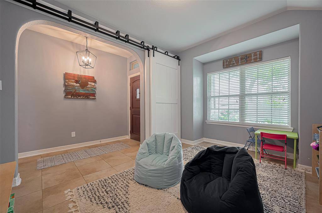 This photo showcases a cozy, well-lit room with a large window and neutral walls. It features a tiled entryway with a modern light fixture and an archway, making it ideal for a playroom or casual living area.