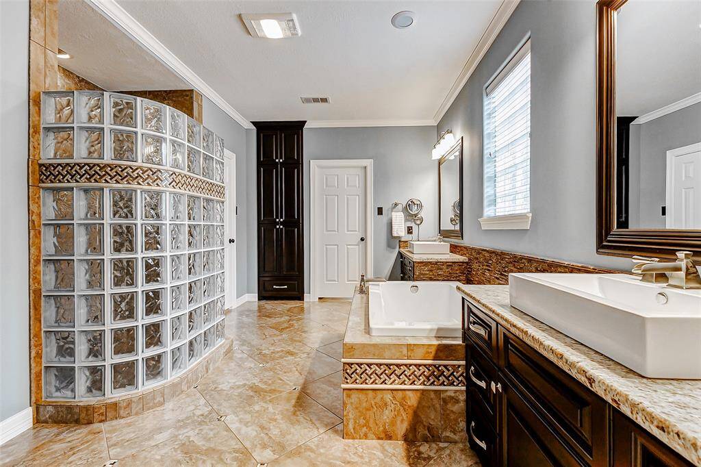 The primary bathroom featuring a curved glass block shower, dual sinks with granite countertops, and a large bathtub.