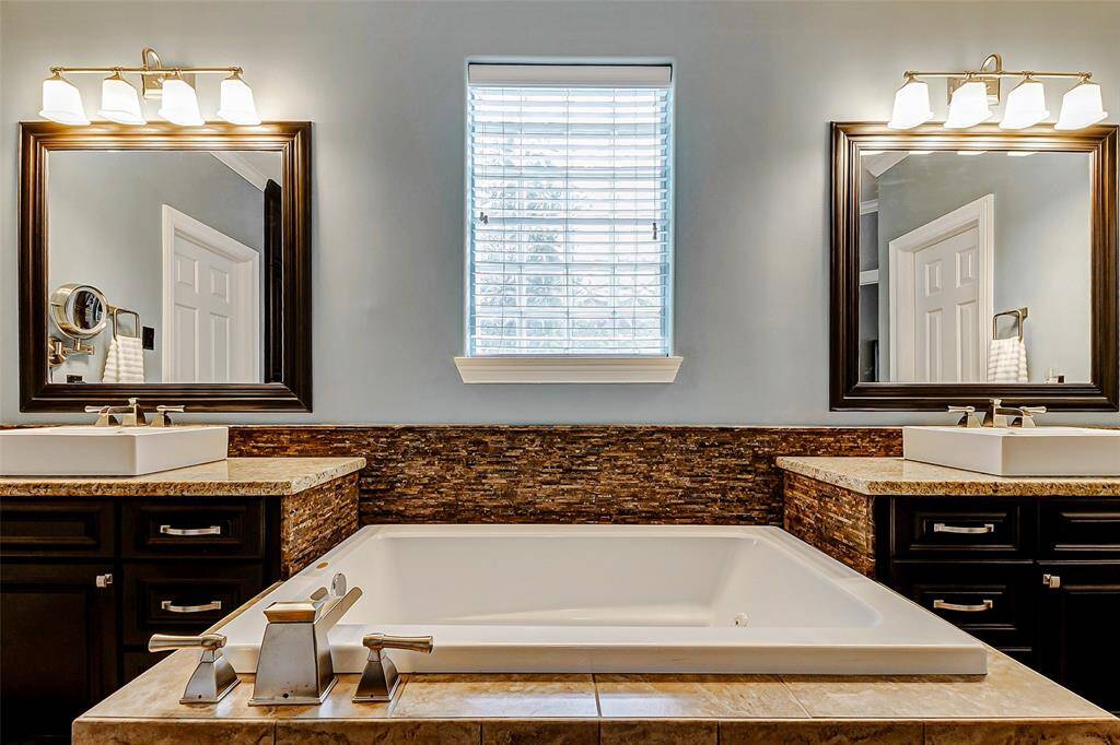 This bathroom features a luxurious soaking tub set between two vanities with modern sinks. Each vanity has a large framed mirror and elegant lighting. The space is accented with a stylish stone backsplash and a central window for natural light.