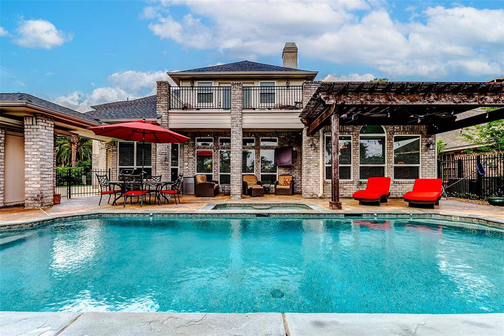 A luxurious backyard with a spacious pool, surrounded by a patio featuring a dining area, lounge chairs, and a pergola.