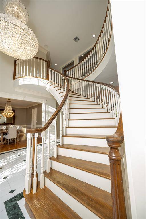 This breathtaking grand staircase gracefully curves upward, featuring rich wood steps, elegant white balusters, and a striking handrail that complements the gleaming marble floors below, all illuminated by a dazzling crystal chandelier.