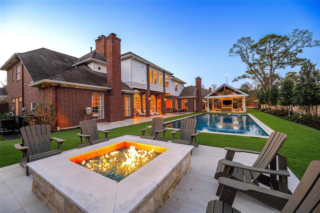 Enhancing the allure of this stunning outdoor retreat, the elegant stone fire pit casts a warm, inviting glow, surrounded by thoughtfully arranged Adirondack chairs, offering the perfect setting for relaxation and entertaining under the open sky.