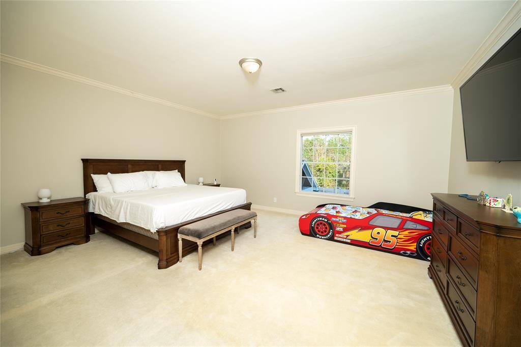 This spacious and inviting bedroom is filled with natural light from a large window, highlighting its soft neutral tones, high ceilings, and well-balanced layout, creating a warm and comfortable retreat.