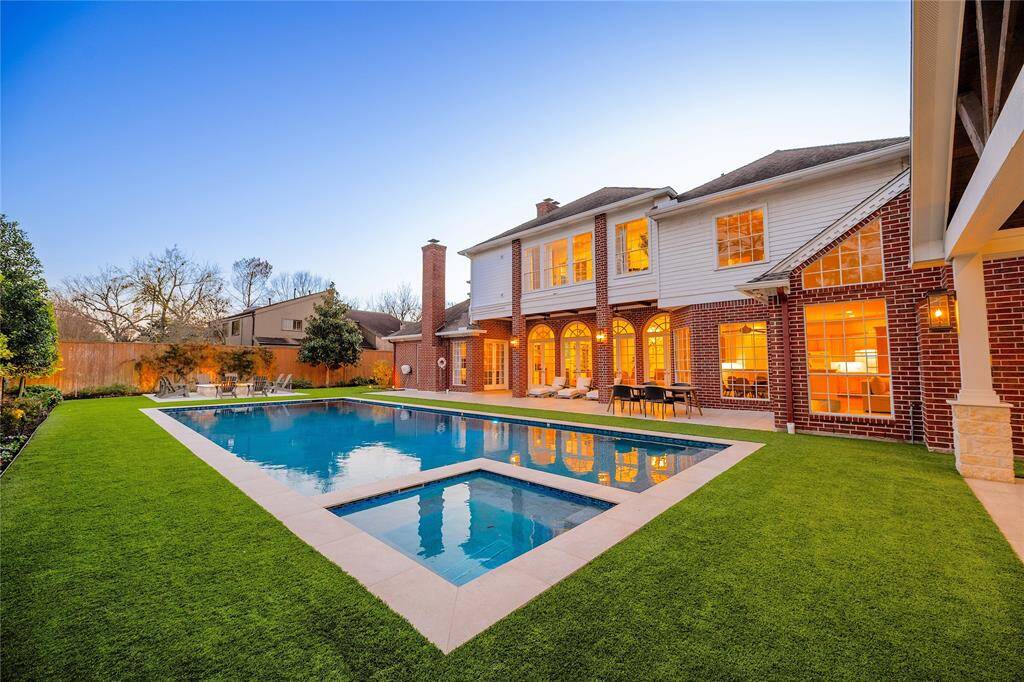 backyard is a stunning retreat featuring a sleek modern pool with a built-in spa, lush green artificial turf, and a spacious covered patio, all set against the backdrop of an elegant, well-lit brick and siding home.