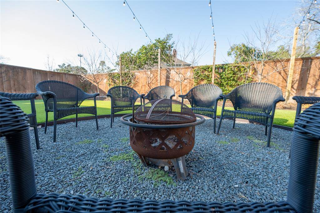 This inviting backyard retreat features a thoughtfully designed fire pit area with a gravel base, surrounded by stylish woven outdoor chairs, ambient string lighting, and a lush backdrop of mature trees and a privacy fence.