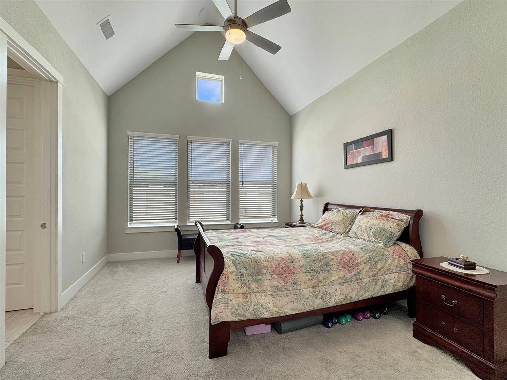The primary bedroom features a vaulted ceiling with a fan, creating an open and airy feel. It has large windows that provide ample natural light, neutral walls, and cozy carpeting.