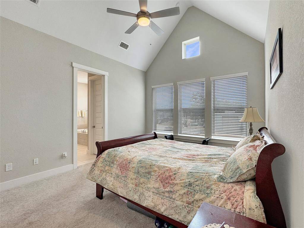 The primary bedroom features a vaulted ceiling with a fan, creating an open and airy feel. It has large windows that provide ample natural light, neutral walls, and cozy carpeting.