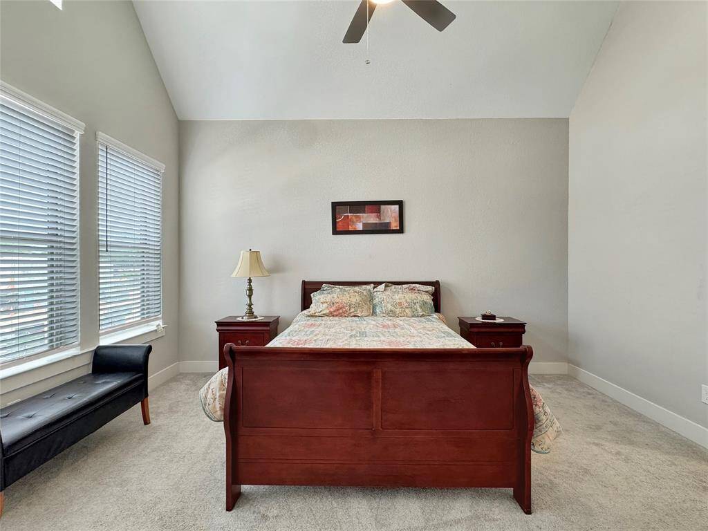 The primary bedroom features a vaulted ceiling with a fan, creating an open and airy feel. It has large windows that provide ample natural light, neutral walls, and cozy carpeting.