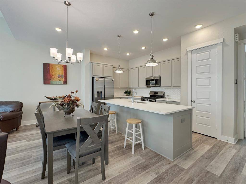 Enjoy a modern kitchen and dining area with light gray cabinetry, stainless steel appliances, and a large island with bar seating. The space features wood-look flooring, recessed lighting, and a stylish chandelier over the dining table, creating a bright and welcoming atmosphere.