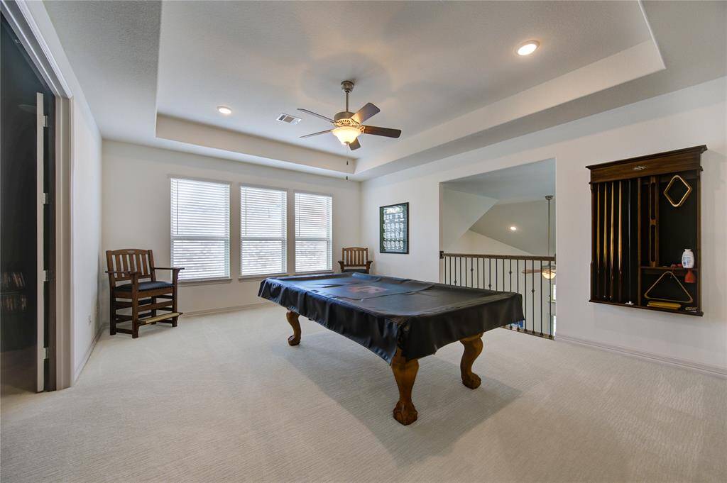 This expansive game room features ample space for a pool table, enhanced by recessed lighting and a ceiling fan. Large windows flood the area with natural light, while a chic railing overlooks the lower level, perfect for entertaining.