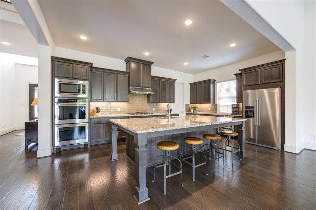 This kitchen features dark wood cabinetry, double ovens, wine refrigerator, stainless steel appliances, and a large granite island with bar seating. The open layout and recessed lighting create a spacious and inviting atmosphere.