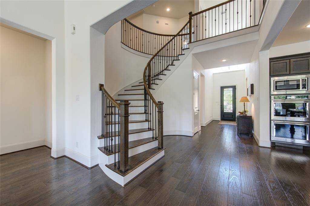 This photo showcases a modern, open-concept entryway with dark hardwood floors and a striking curved staircase with elegant iron railings. The space is bright and airy, with high ceilings and a view into a sleek kitchen area.