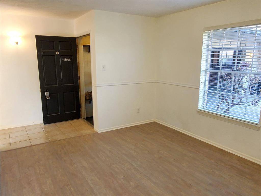 This room features light wood flooring, a window with blinds allowing natural light, and a contrasting dark front door with adjacent tile flooring. The walls are neutral, offering a blank canvas for personalization.