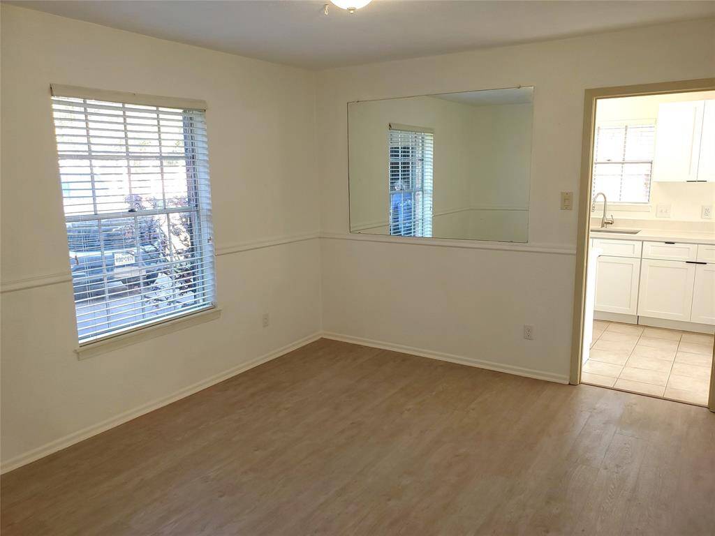 This photo shows a bright room with light wood flooring, a large window with blinds, and a mirrored wall. It opens into a kitchen featuring white cabinetry and tiled flooring.