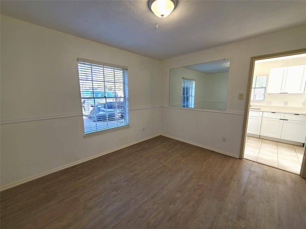 This room features a bright, open space with wood-style flooring and a large window for natural light. There's a wall mirror enhancing the sense of space and a doorway leading to a kitchen with white cabinetry.