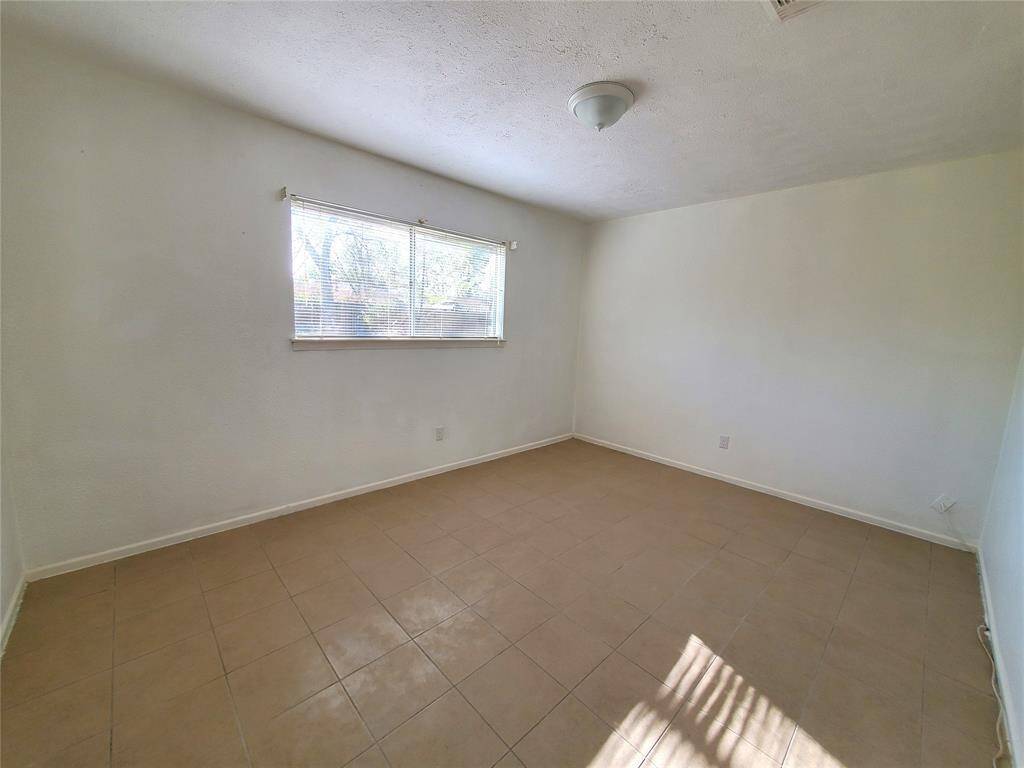 This room features neutral tile flooring, light-colored walls, and a ceiling light fixture. A window with blinds provides natural light, creating a bright and airy space. Ideal for a bedroom or office.
