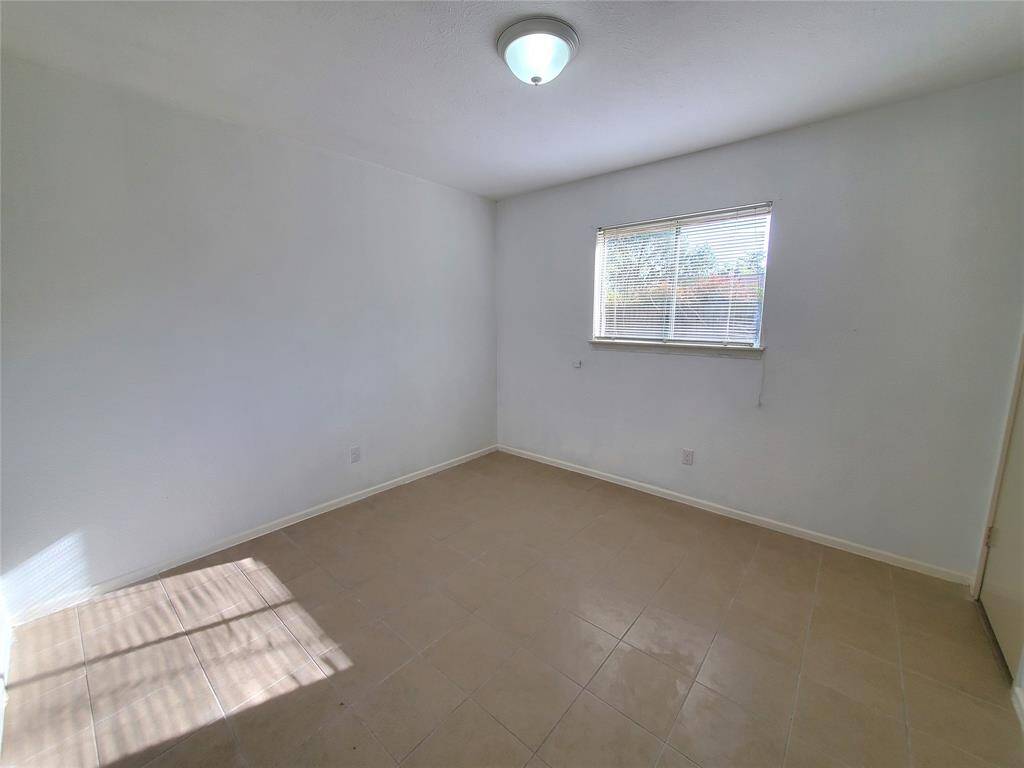 A bright, empty room with light-colored tiled flooring and white walls. It has a window with blinds allowing natural light to fill the space, and a ceiling light fixture. Ideal for customization.