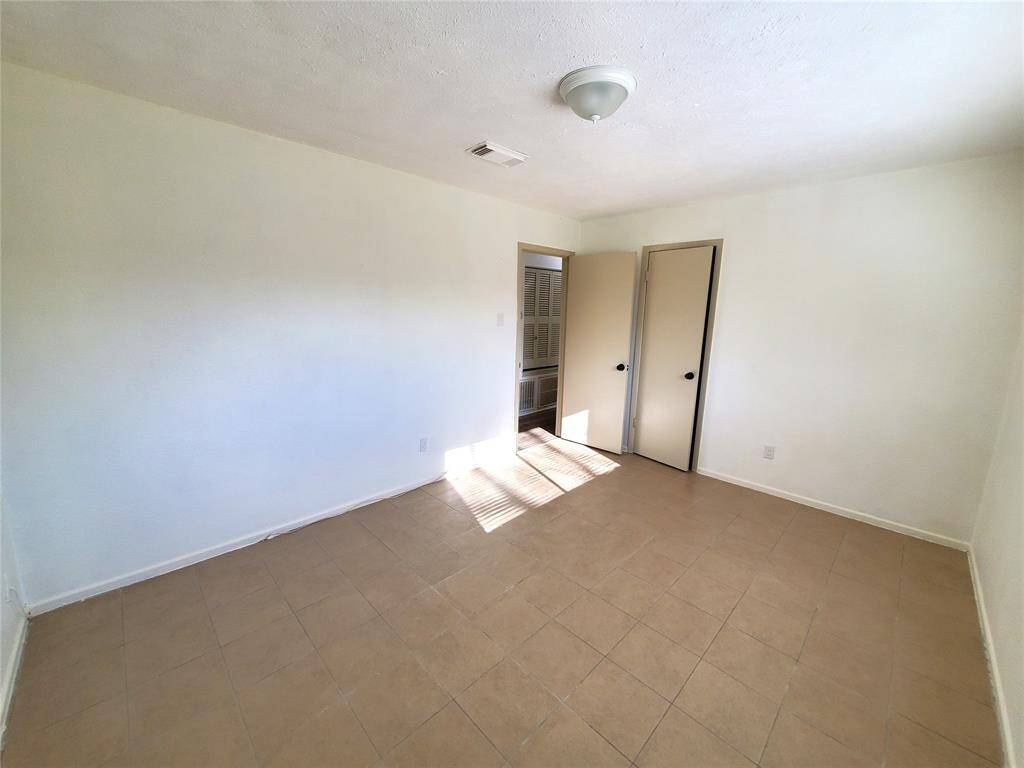 This is a bright, empty room with neutral-colored tile flooring and white walls. It features a ceiling light fixture and two doors, one slightly ajar, leading to additional spaces. Natural light streams in from an unseen window, creating a welcoming atmosphere.