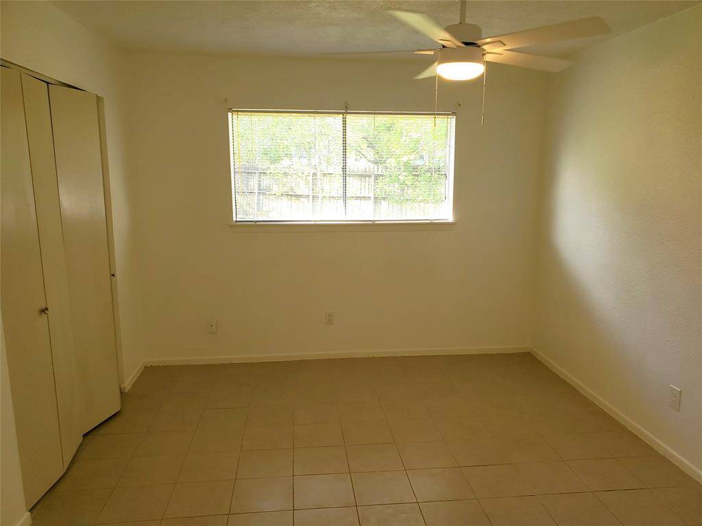 This room features neutral tile flooring, a ceiling fan for comfort, a large window providing natural light, and a closet with double doors, offering a clean and versatile space.