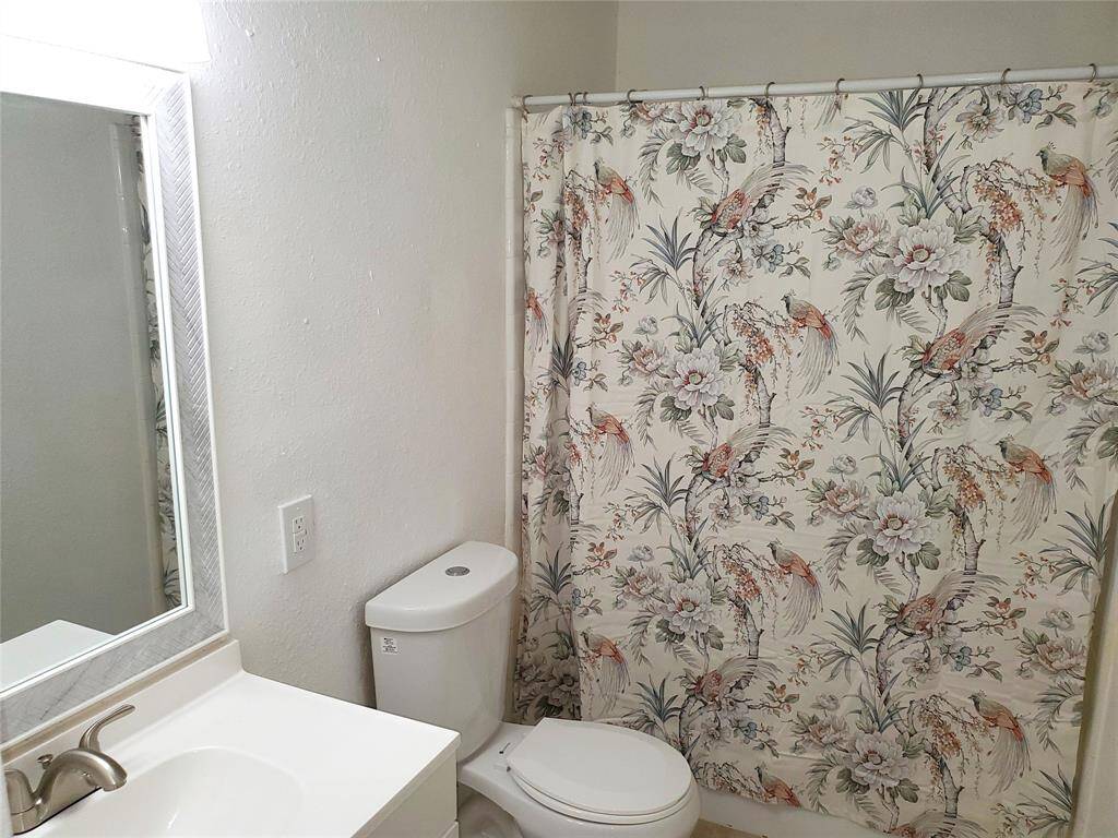 This bathroom features a clean, white design with a floral and bird-patterned shower curtain. It includes a standard toilet, a sleek vanity with a modern faucet, and a large mirror, creating a bright and inviting space.