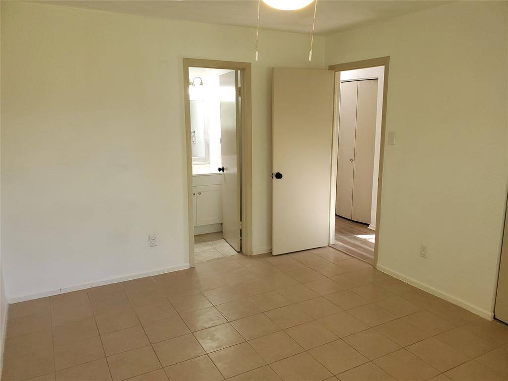 This photo shows a bright, empty room with tiled flooring and light-colored walls. It features two open doors leading to a bathroom and a hallway, providing a clean and versatile space ready for personal touches.