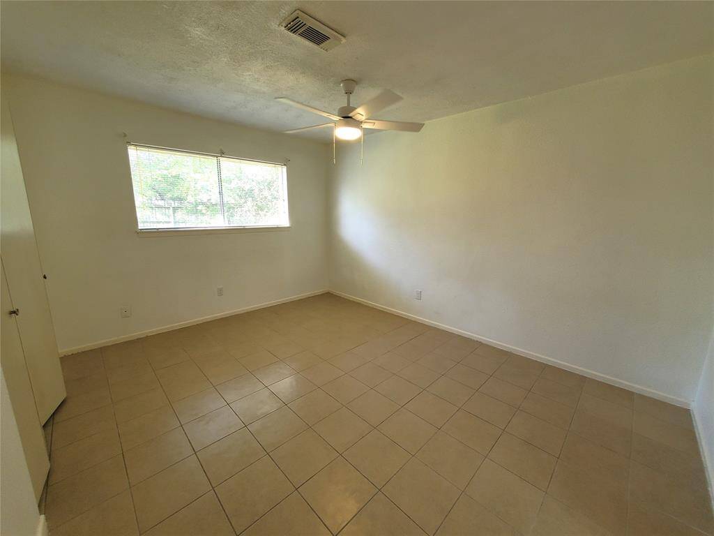 This room features neutral-colored tile flooring, a ceiling fan, and a large window that provides natural light. The walls are plain, offering a blank canvas for personalization.