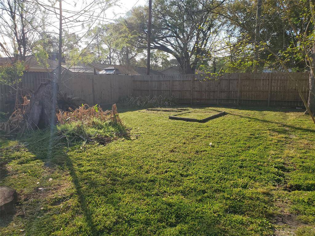 This photo shows a spacious backyard with a wooden fence, surrounded by tall trees offering shade. The area is mostly grassy, with a small garden bed and a tree stump, providing a good space for outdoor activities or gardening.