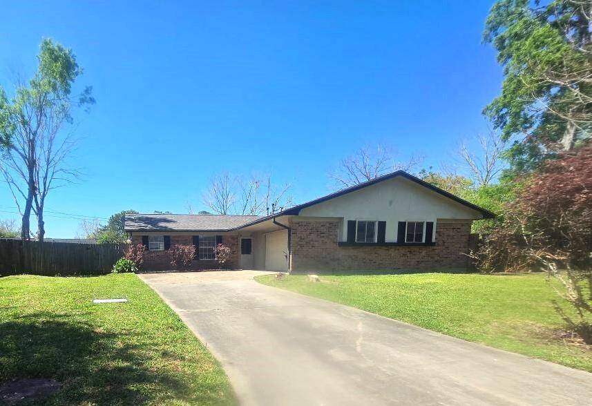 Charming single-story brick home with a sloping roof and attached garage. It features a spacious driveway and a well-maintained front lawn, surrounded by mature trees for added privacy.