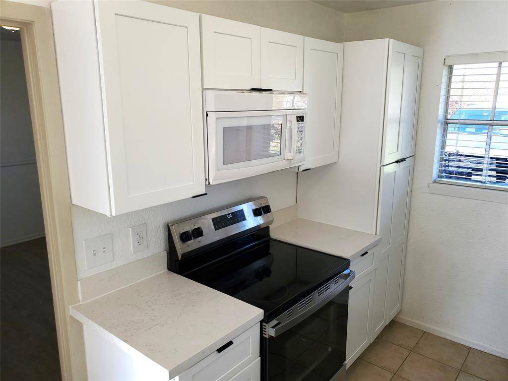 Compact kitchen with modern white cabinetry, an electric stove, and a built-in microwave. Light flows in through a window with blinds, offering a clean and bright space.
