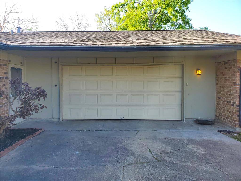 This photo shows a single-car garage with a light beige door, surrounded by brick and siding. There's a small outdoor light fixture on the right, a driveway with some cracks, and a small shrub on the left.