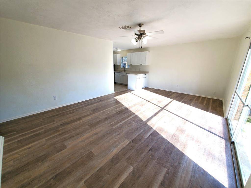 Bright and spacious open-plan living area with wood flooring, ceiling fan, and large windows for natural light. The kitchen features white cabinets and a functional layout.