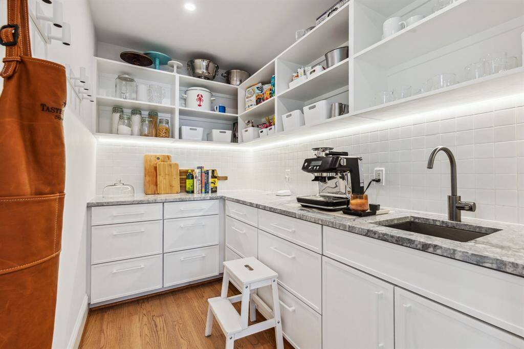 Enjoy plenty of room for small appliances and staples in this beautifully arranged walk-in storage pantry, where cabinetry and shelving are trimmed with tile backsplashes, Super White Calacatta Dolomite counters, under-cabinet lighting and a sink.