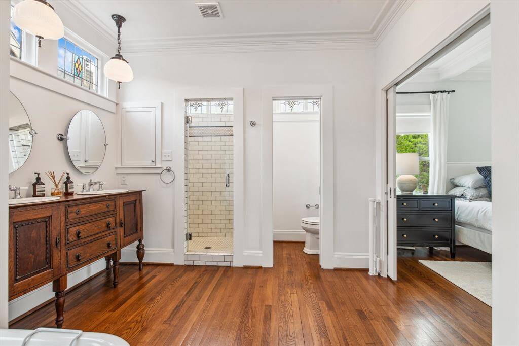 The breathtaking primary bathroom is anchored by an antique furniture-style double vanity accented by lovely pendant lighting, two medicine cabinets, stained glass windows and tilted round mirrors.