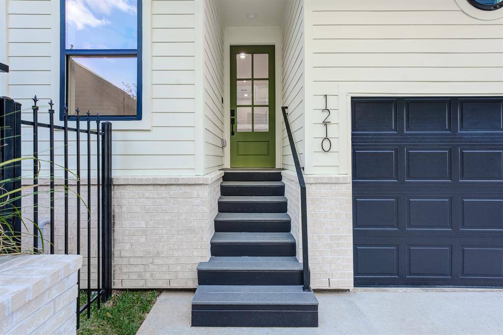 This photo showcases a modern stairway entrance with a stylish green door, black accented double paned windows, adjacent to a matching garage door. The exterior features Hardie Plank siding and neutral bricks, and a small landscaped area with shrubs.